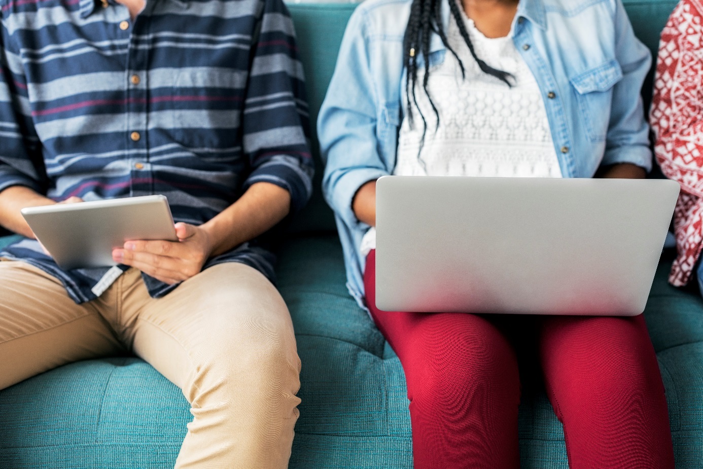 Junger Mann mit Tablet in der Hand und junge Frau mit Laptop auf dem Schoß sitzen nebeneinander.