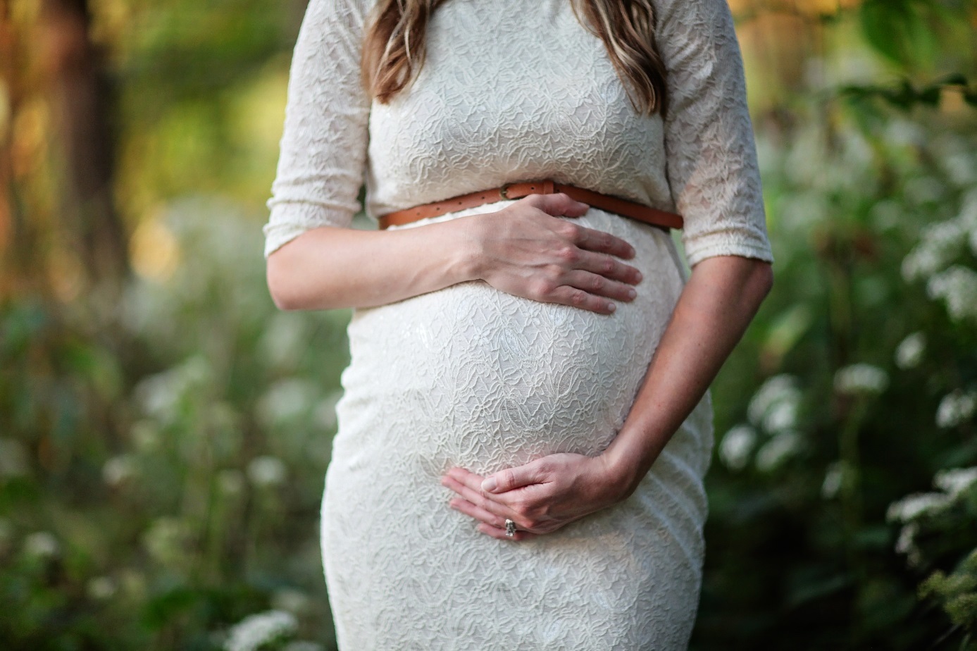 Schwangere in weißem Kleid hält Hände vor dem Bauch. 