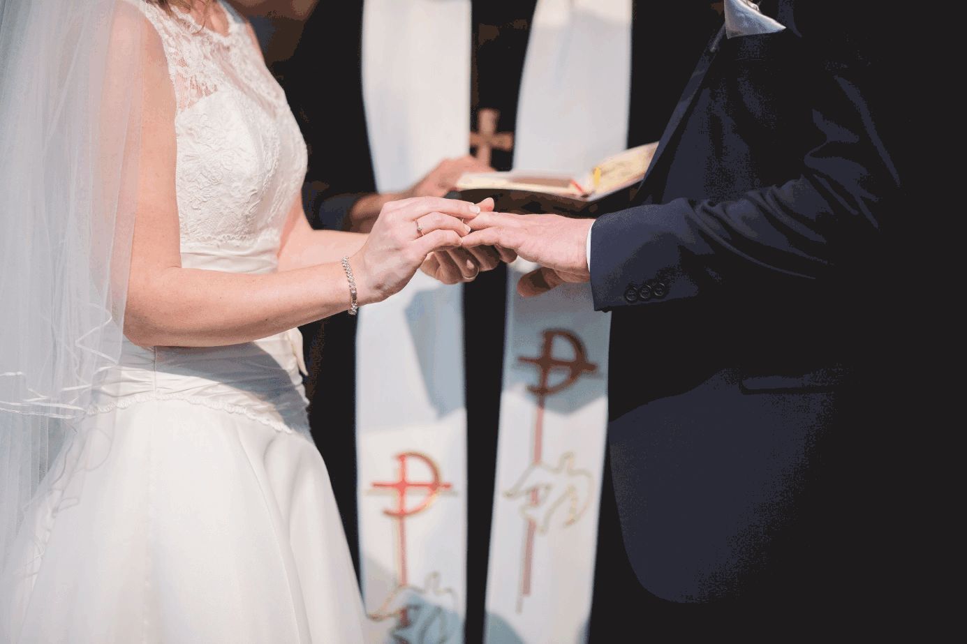 Braut steckt Bräutigam den Ring auf, im Hintergrund steht der Priester.